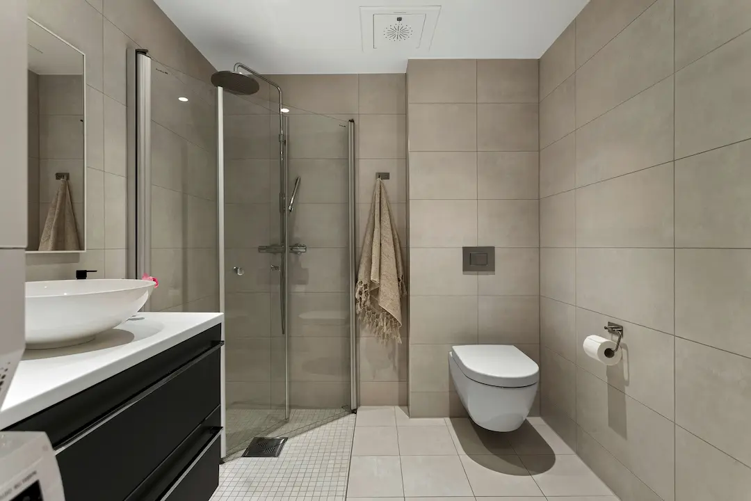 Nowoczesna, minimalistyczna łazienka z beżowymi płytkami na ścianach, szklanym prysznicem typu walk-in, podwieszaną toaletą, białą umywalką nablatową i czarną szafką podumywalkową.